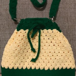 May include: A handmade crochet bag with a drawstring closure. The bag features a cream-colored body with a green base, trim, and straps. The bag is designed with a textured stitch pattern and has a green drawstring.