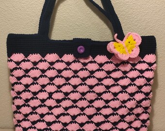 May include: A handmade crochet tote bag with a navy blue base and handles. The body of the bag features a pink shell stitch pattern. A yellow and pink butterfly adorns the front, and a purple button secures the top.