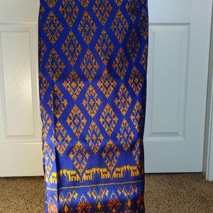 May include: A long, blue fabric with a repeating diamond pattern in yellow and orange. The fabric is displayed on a white stand. The pattern is consistent across the length of the fabric.
