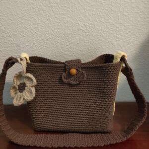 May include: A handmade brown crochet bag with a flower accent. The bag features a rectangular shape with a strap and a wooden button closure. The flower accent is a lighter shade of brown. The bag is sitting on a wooden surface.