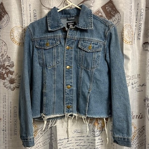 May include: A light blue denim jacket with a frayed hem. The jacket has a classic collar, button-down front, and two chest pockets. The brand "boohoo" is visible on the label. The jacket is hanging on a white hanger.