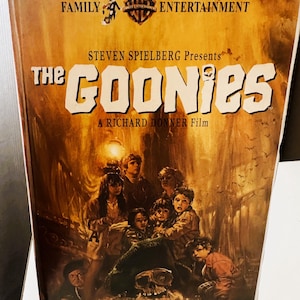 Könnte beinhalten: Eine VHS-Kassette des Films "Die Goonies" mit dem Warner Bros. Entertainment-Logo. Das Cover zeigt eine Gruppe von Kindern in einer Schatzhöhle, mit dem Filmtitel in weißen Buchstaben.
