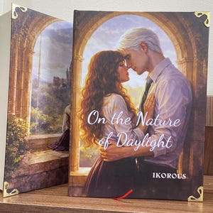 May include: Two hardcover books with gold corner accents. The book on the right features a couple embracing, with the title "On the Nature of Daylight" and the author's name "IKOROUS". The spine of the book on the left shows a landscape painting.