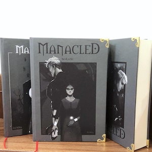 May include: Three hardcover books with the title "MANACLED" on the cover. The covers are gray with a black and white illustration of two people. The corners of the books are gold. The books are on a wooden shelf.