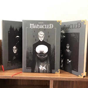 May include: Three hardcover books with the title "Manacled" are displayed on a wooden shelf. The book covers are primarily gray with illustrations of figures and the title in white. Gold corner protectors add a decorative touch.