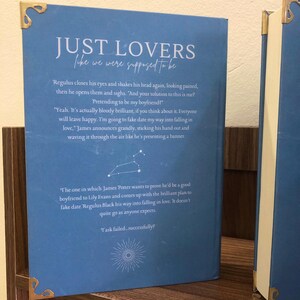 Pu&ograve; includere: Un libro blu intitolato "JUST LOVERS" con il sottotitolo "the we were supposed to be." La copertina presenta estratti di testo e un grafico di costellazione. Gli angoli dorati aggiungono un tocco decorativo.