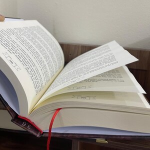 May include: An open book with a red ribbon bookmark. The book has a brown leather cover and pages with printed text. Page numbers 198, 201, and 203 are visible. The book is on a wooden surface.