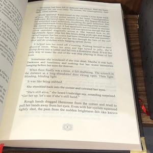 May include: Open book with text on the pages. The text is in English and describes a scene with the character Hermione. The book has a dark brown cover and a red ribbon bookmark.