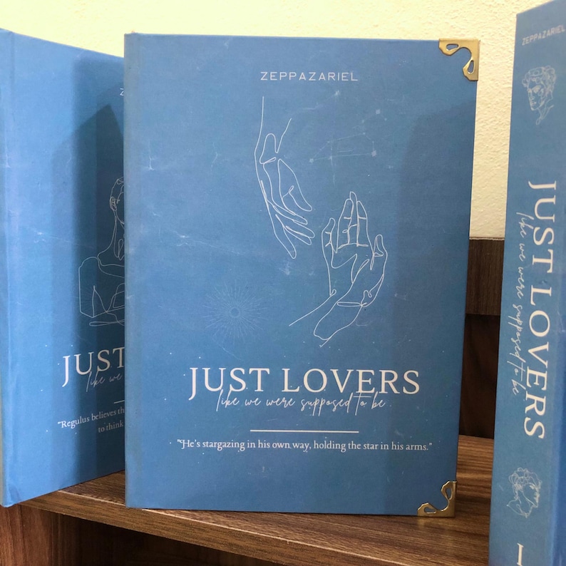 Pu&ograve; includere: Una pila di libri con copertine blu. Il libro principale &egrave; intitolato "JUST LOVERS" con il sottotitolo "like we were supposed to be." La copertina presenta un disegno a tratto bianco di mani e il nome dell'autore, ZEPPAZARIEL.