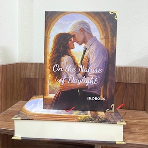 May include: A hardcover book titled "On the Nature of Daylight" with a romantic cover illustration of a couple embracing. The book is resting on another book with a decorative cover. The author's name, IKOROUS, is visible.