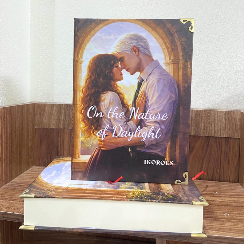 May include: A hardcover book titled "On the Nature of Daylight" with a romantic cover illustration of a couple embracing. The book is resting on another book with a decorative cover. The author's name, IKOROUS, is visible.