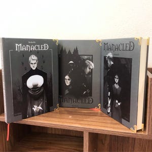 May include: Three hardcover books titled "Manacled" are displayed on a wooden shelf. The book covers are dark gray with illustrations of characters and the title in white. Gold corner protectors and red bookmarks are visible.