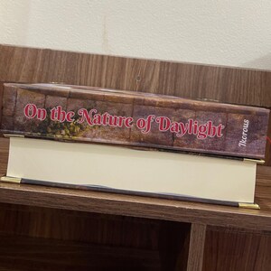 May include: A hardcover book titled "On the Nature of Daylight" with a brown cover and gold-colored corner accents. The book is resting on a shelf. The author's name, Ikorous, is printed on the side.