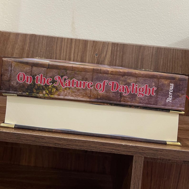 May include: A hardcover book titled "On the Nature of Daylight" with a brown cover and gold-colored corner accents. The book is resting on a shelf. The author's name, Ikorous, is printed on the side.