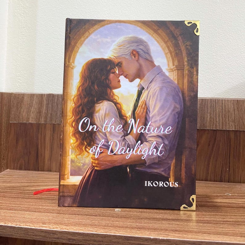 May include: A hardcover book titled "On the Nature of Daylight" with a romantic illustration of a couple embracing. The book cover features a warm color palette with gold corner accents and the author's name, IKOROUS, at the bottom.