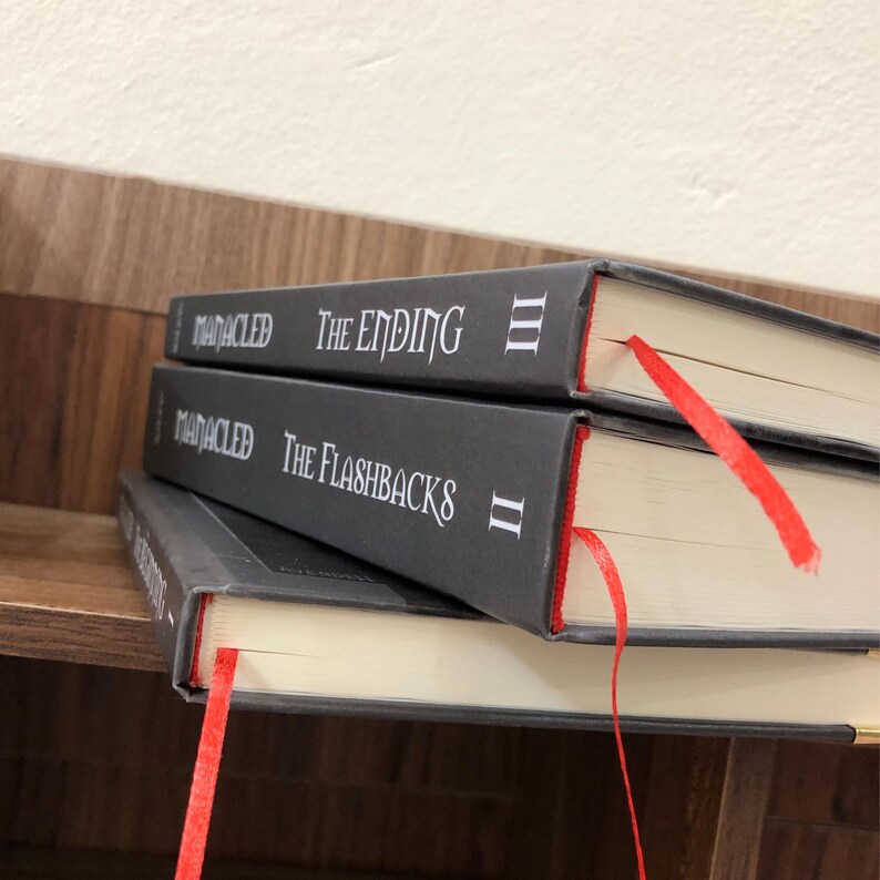 May include: Three hardcover books stacked on a wooden shelf. The books have dark gray covers with white text that reads "MANACLED THE ENDING III" and "MANACLED THE FLASHBACKS II". Each book has a red ribbon bookmark.