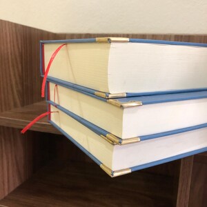 Pu&ograve; includere: Tre libri con copertina rigida impilati su una mensola di legno. I libri hanno copertine azzurre, angoli dorati e segnalibri a nastro rosso. Le pagine sono color crema.
