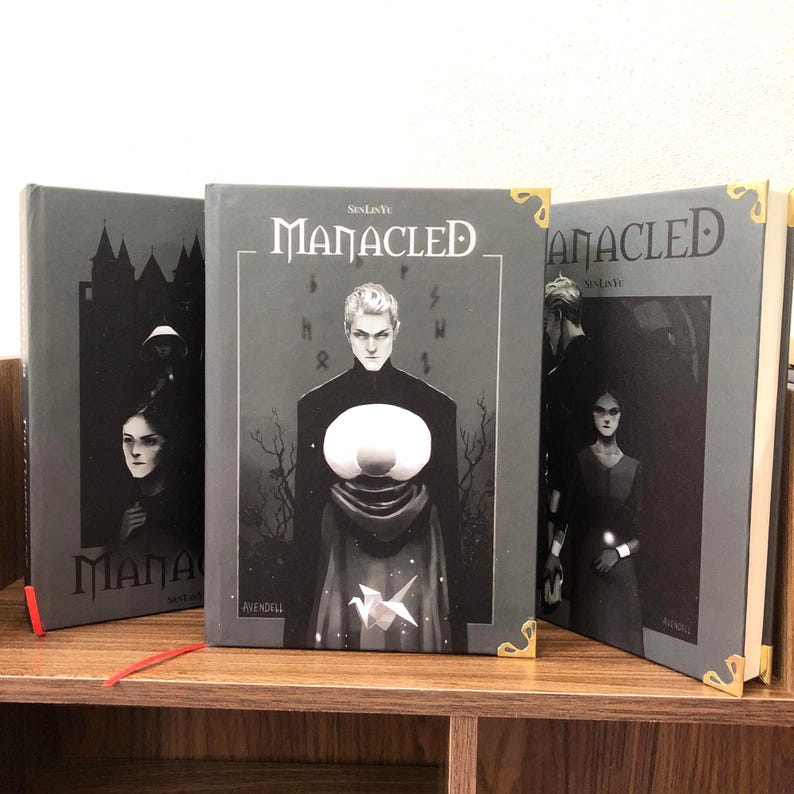 May include: Three hardcover books with the title "Manacled" are displayed on a wooden shelf. The book covers are primarily gray with illustrations of figures and the title in white. Gold corner protectors add a decorative touch.