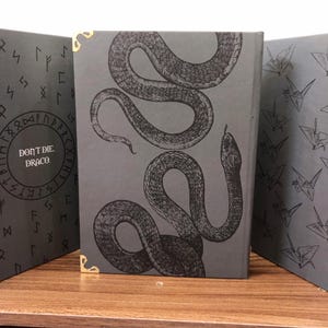 May include: A set of three dark gray books with gold corner protectors. The center book features a detailed illustration of a snake. The left book has the text "DON'T DIE. DRACO." in a circle. The right book has a pattern of origami cranes.