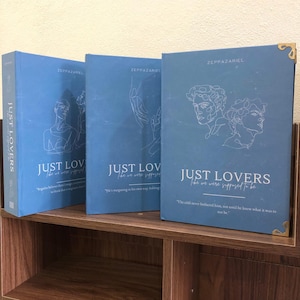 Pu&ograve; includere: Tre libri blu con copertina rigida con il titolo "JUST LOVERS" e il nome dell'autore "ZEPPAZARIEL" stampati sulla copertina. Le copertine presentano illustrazioni a tratteggio di figure e citazioni.