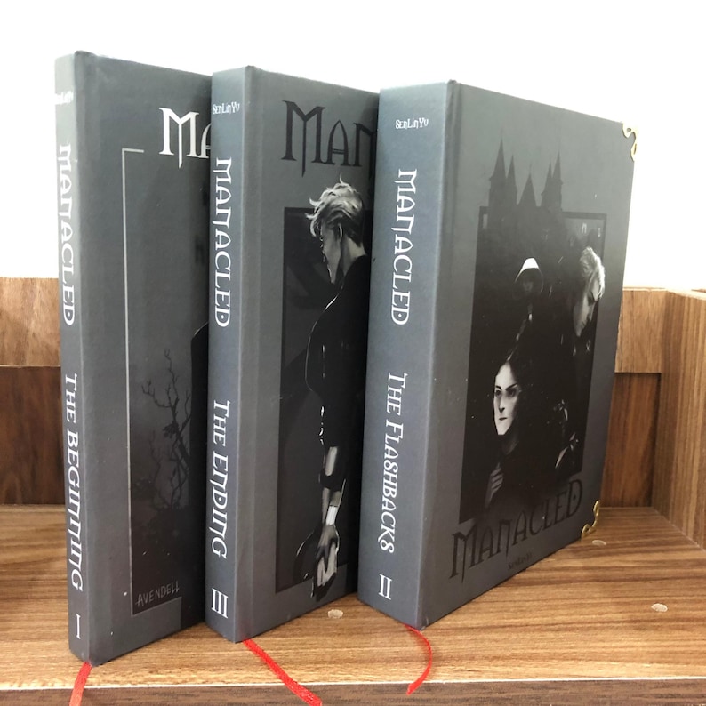 May include: Three hardback books with gray covers are displayed on a wooden shelf. The titles are "MANACLED: The Beginning", "MANACLED: The Ending", and "MANACLED: The Flashbacks". Each book has a different illustration and a red ribbon bookmark.