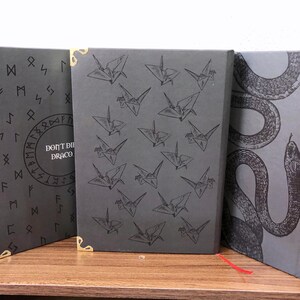 May include: Three hardback books with dark gray covers. The center book features a pattern of origami cranes. The left book has runes and the text "DON'T DIE DRACO." The right book has a snake illustration. Gold corner protectors are visible.