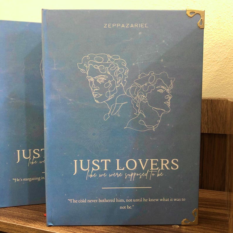 Pu&ograve; includere: Un libro blu intitolato "JUST LOVERS" con il sottotitolo "like we were supposed to be." La copertina presenta ritratti a linea bianca e il nome dell'autore, ZEPPAZARIEL. Gli angoli dorati aggiungono un tocco decorativo.