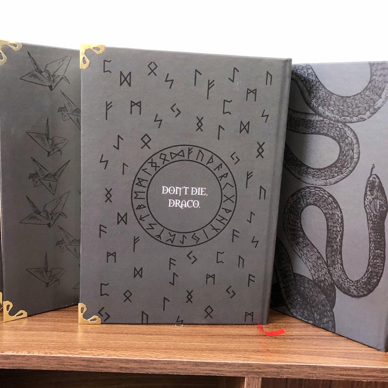 May include: A collection of three hardback books with dark gray covers. The center book features the text "DON'T DIE. DRACO." within a circular design, surrounded by runes. The other books have origami and snake illustrations.