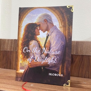 May include: A hardcover book titled "On the Nature of Daylight" featuring a romantic illustration of a couple embracing. The cover art showcases a man and woman in a warm, sunlit setting. The author's name, IKOROUS, is at the bottom.