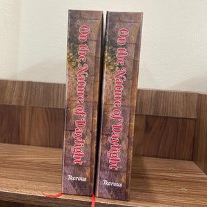 May include: Two books titled "On the Nature of Daylight" are displayed upright on a wooden shelf. The book spines feature a brick wall design with the title in red script and the author's name, "Ikorous," at the bottom. A red bookmark is visible.