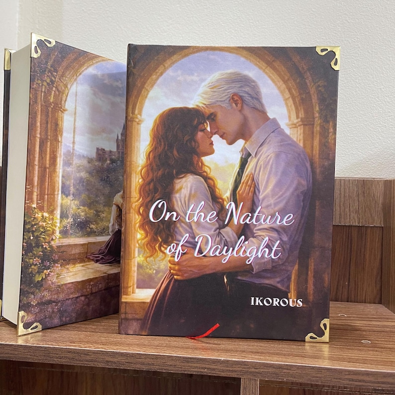 May include: Two hardcover books with gold corner accents. The book on the right features a couple embracing, with the title "On the Nature of Daylight" and the author's name "IKOROUS". The spine of the book on the left shows a landscape painting.