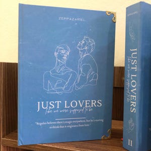 Pu&ograve; includere: Un libro blu intitolato "JUST LOVERS like we were supposed to be" di Zeppazariel. La copertina presenta un disegno a tratto di due figure e una citazione. &Egrave; visibile anche il dorso del libro, con il titolo e il nome dell'autore.