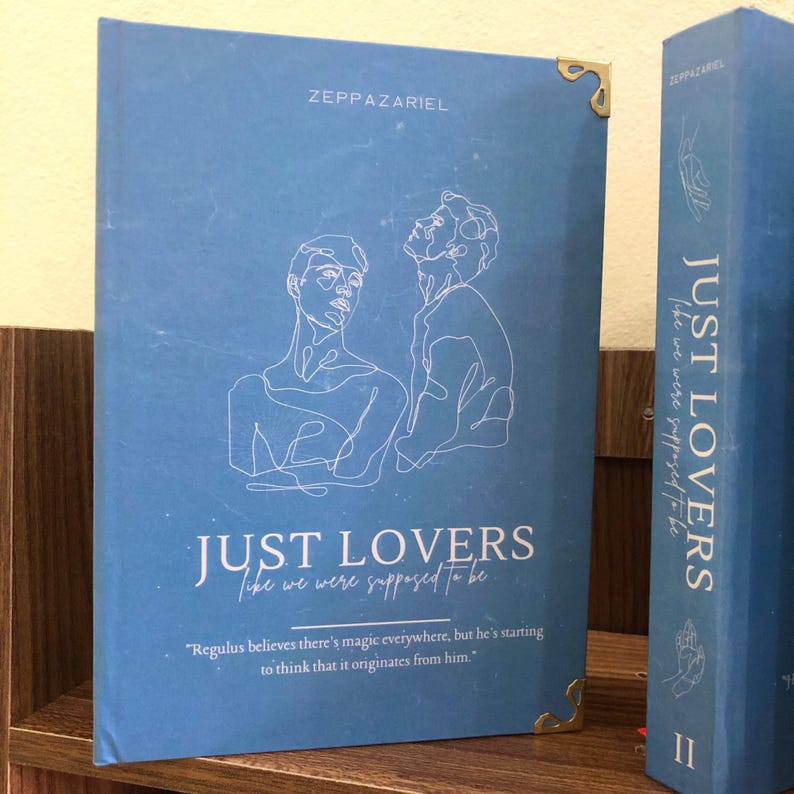 Pu&ograve; includere: Un libro blu intitolato "JUST LOVERS like we were supposed to be" di Zeppazariel. La copertina presenta un disegno a tratto di due figure e una citazione. &Egrave; visibile anche il dorso del libro, con il titolo e il nome dell'autore.