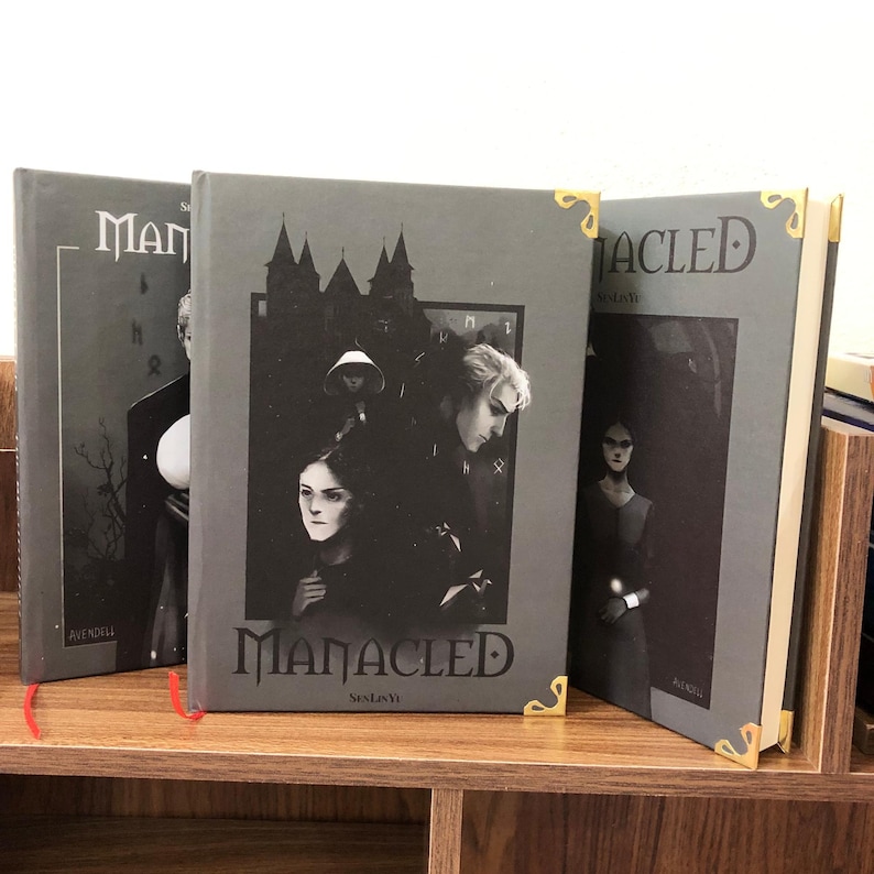 May include: Three hardback books with the title "MANACLED" in a gothic font. The covers are gray with illustrations of figures and a castle. Gold corner protectors and red bookmarks add detail.