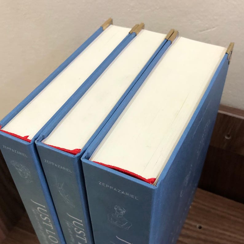 Pu&ograve; includere: Tre libri con copertina rigida con copertine azzurre e pagine bianche. I dorsi sono blu con il titolo "Just Love" e il nome dell'autore. Ogni libro ha un segnalibro a nastro rosso e angoli dorati.