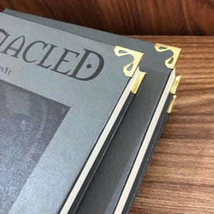 May include: Close-up of a stack of books with gray covers and gold corner protectors. The book titles are partially visible, with the word "ACLE" in large, dark letters. The edges of the books are white, and the background is a wooden surface.