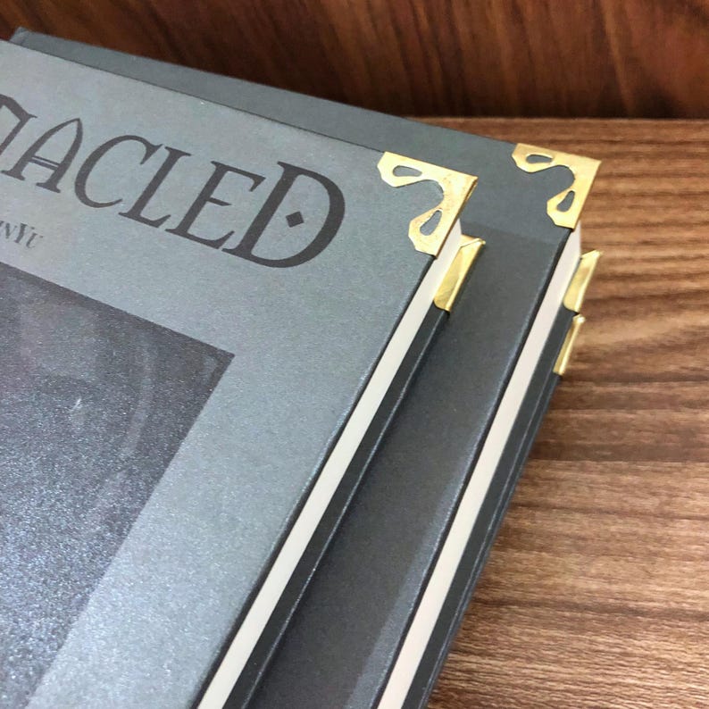 May include: Close-up of a stack of books with gray covers and gold corner protectors. The book titles are partially visible, with the word "ACLE" in large, dark letters. The edges of the books are white, and the background is a wooden surface.