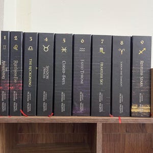 May include: A collection of nine hardcover books with black covers, each featuring a gold astrological symbol and title. The spines display the series title, author's names, and volume numbers. The books are arranged on a wooden shelf.