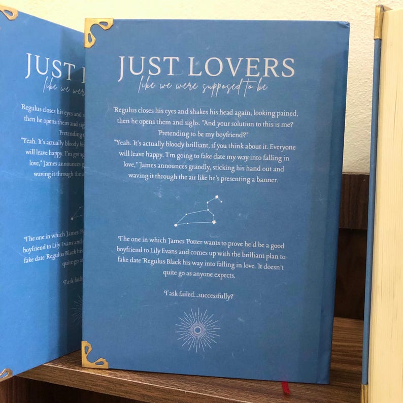 Pu&ograve; includere: Un libro blu intitolato "JUST LOVERS like we were supposed to be" con dettagli dorati agli angoli. La copertina presenta estratti di testo e un riassunto della trama. Il dorso del libro &egrave; visibile.