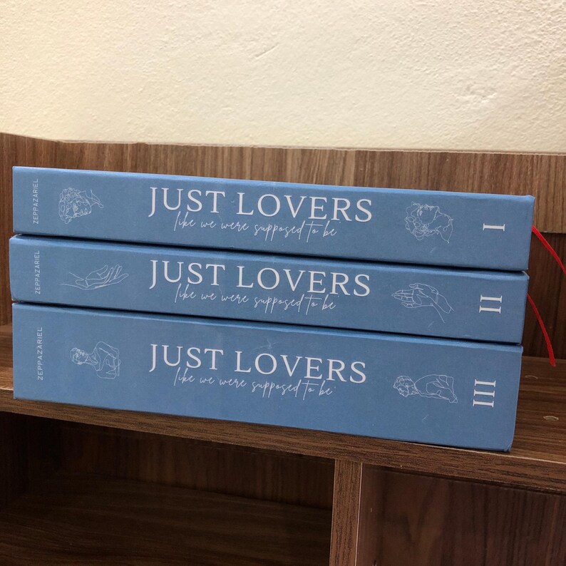 Pu&ograve; includere: Tre libri impilati con copertine azzurre. Il titolo "JUST LOVERS" e il sottotitolo "like we were supposed to be" sono stampati in bianco. I numeri romani I, II e III sono visibili sui dorsi.