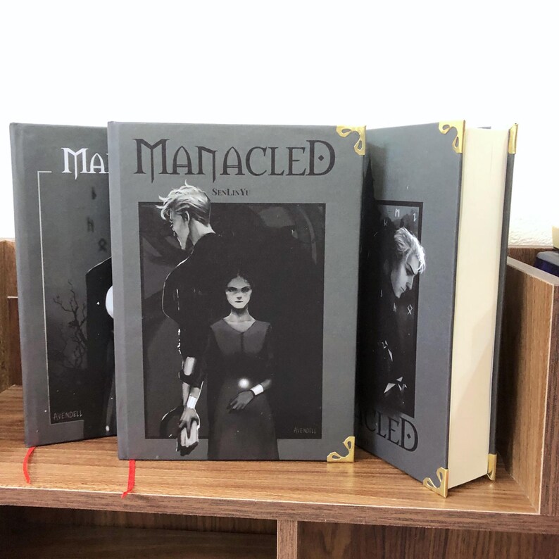 May include: Three hardcover books with the title "MANACLED" on the cover. The covers are gray with a black and white illustration of two people. The corners of the books are gold. The books are on a wooden shelf.