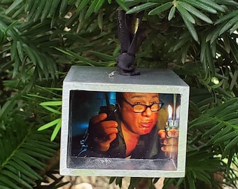 May include: A silver metal ornament with a clear plastic window showing a photo of a person wearing glasses and holding a lighter. The ornament is hanging from a black cord.