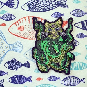 May include: A green and yellow embroidered patch of a cat wearing an octopus costume. The patch is on a white background with a pattern of blue and orange fish.