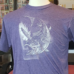 May include: Purple short-sleeve t-shirt featuring a white illustration of a fish with a horn. The fish has an open mouth and detailed scales. The shirt is displayed on a mannequin. The background includes artwork and a press.