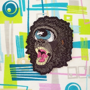 May include: A black and gray embroidered patch of a gorilla with a large blue eye and an open mouth showing teeth.