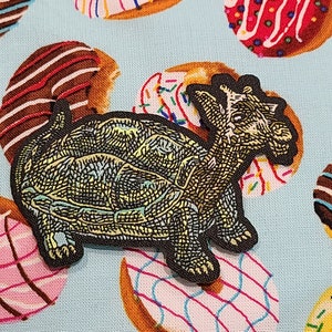 May include: A black and green embroidered patch of a turtle with a detailed shell and a long neck. The patch is on a blue fabric background with a pattern of colorful donuts.