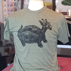 Op de afbeelding: Een groen T-shirt met een zwart-witte grafische print van een wezen dat gedeeltelijk giraffe en gedeeltelijk schildpad is.