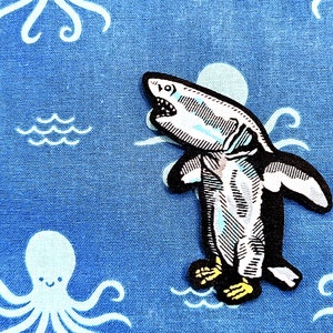 May include: A black and white embroidered patch of a shark wearing a penguin costume. The patch is on a blue fabric background with white octopus designs.
