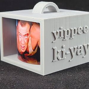 May include: A gray 3D printed box with a handle, featuring a picture of Bruce Willis from the movie Die Hard, and the text "yippee ki-yay" on the side.