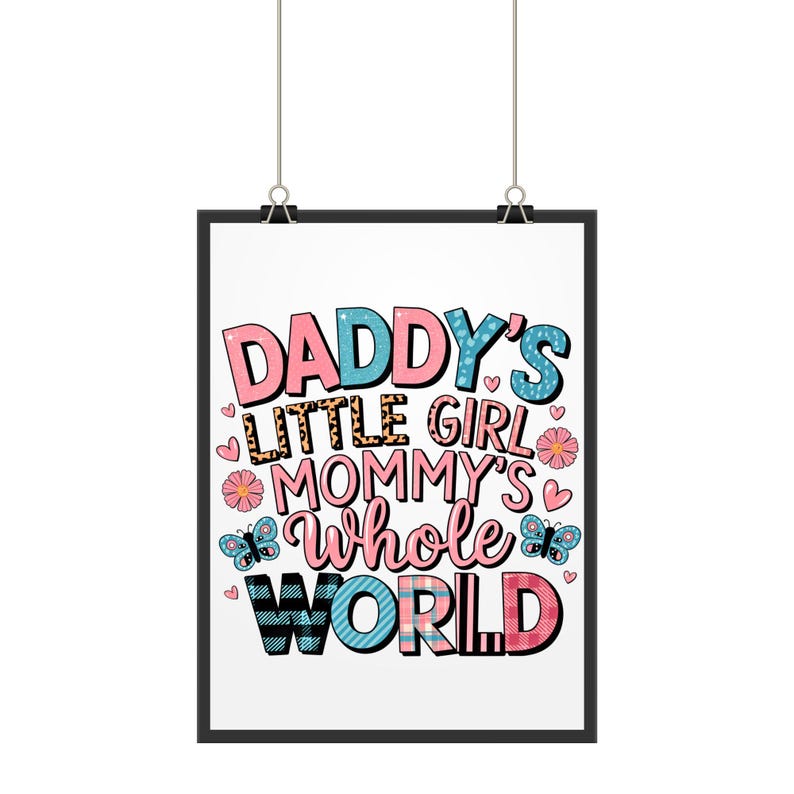 以下が含まれることがあります： ピンク、ブルー、ブラックの文字で「Daddy's Little Girl Mommy's Whole World」と書かれた、黒い額縁のプリント。ハート、蝶、花のパターンが背景になっている。